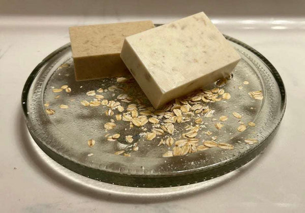 Oatmeal Scrub Soapbar with two beige rectangular bars, one speckled with oats, beside scattered oat flakes